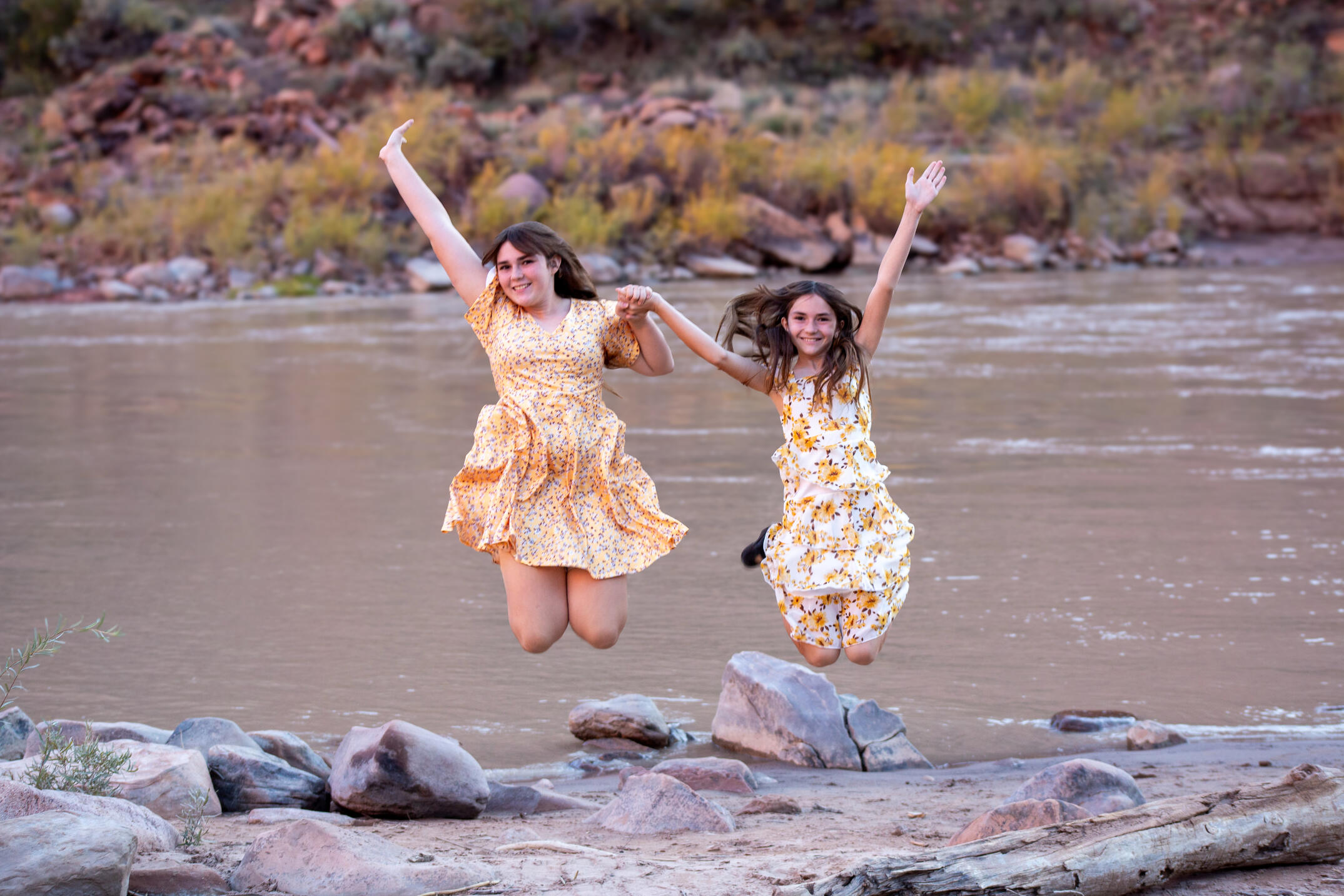 two sisters in yellow dresses hold hand and jump up in front of the Colorado river family photos
