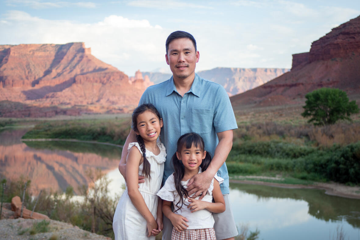 dad with two daughters standing in front of river and large expanse of moab canyon walls behind them family photos