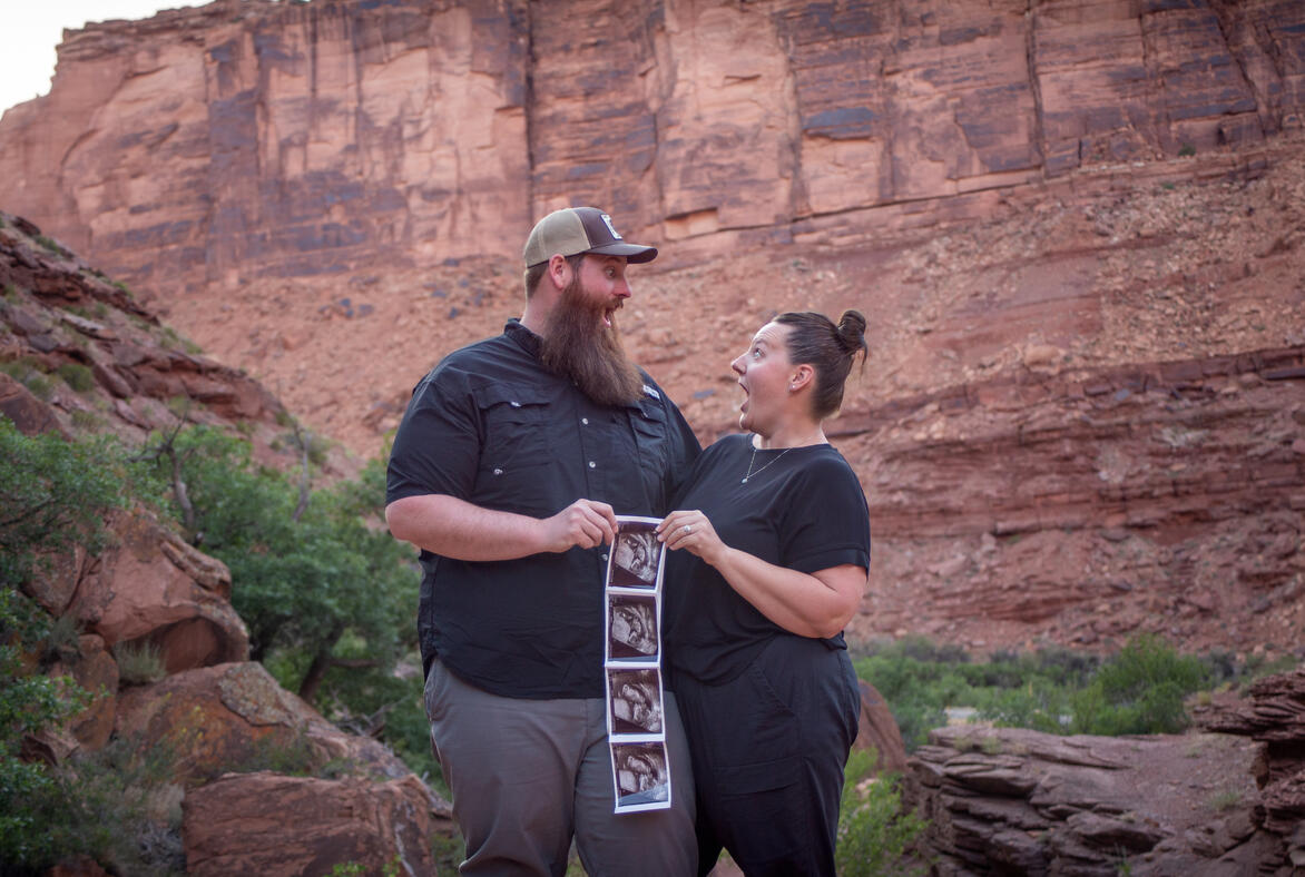 mom and dad to be looking at each other with surprised faces while holding ultrasound pictures red rock canyon wall behind baby announcement maternity