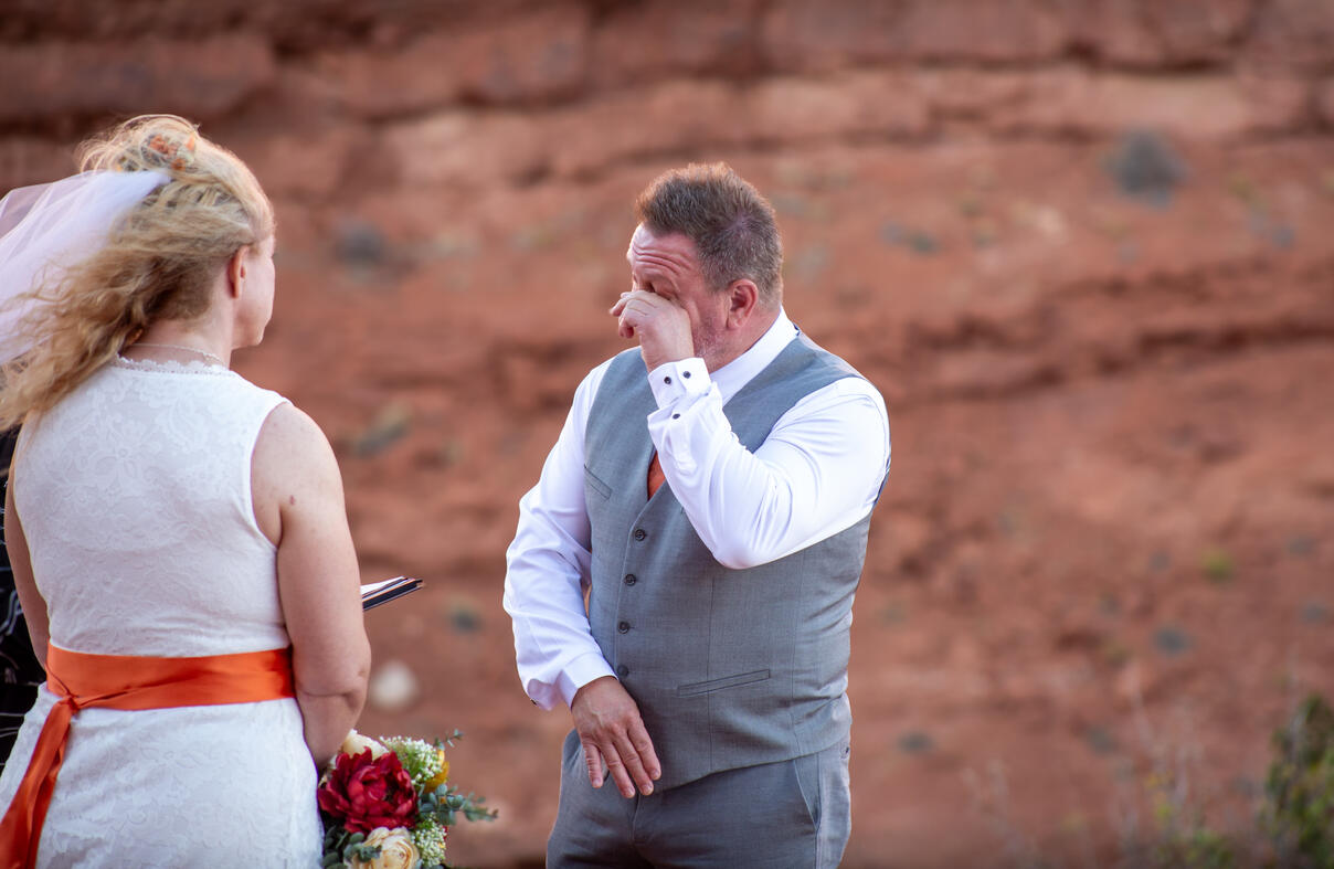 soon to be husband wipes his eyes during ceremony can see soon to be wife from her back as she looks on red rocks behind them small wedding marriage