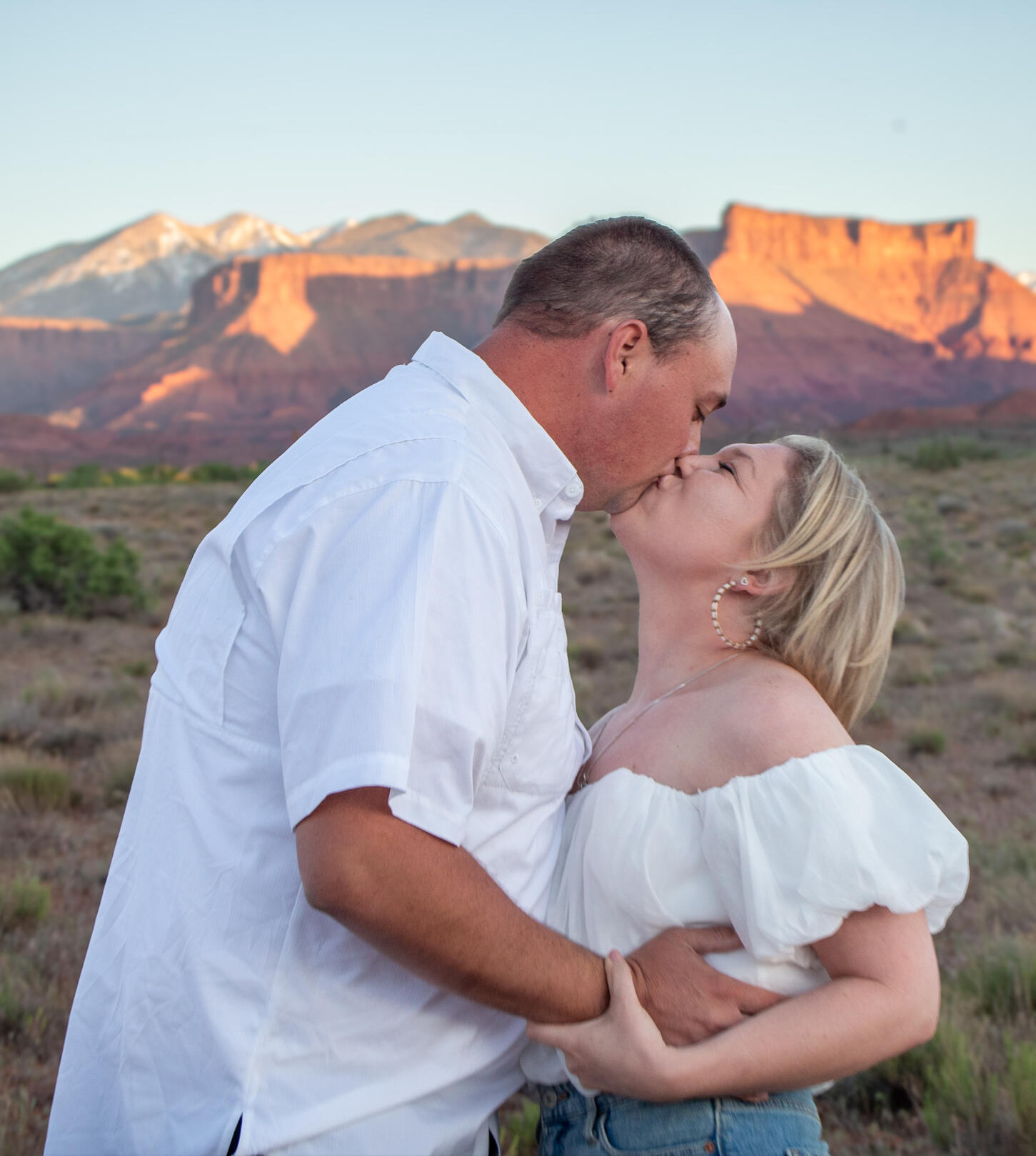 engaged couple kiss with red rocks and mountains behind proposal moab