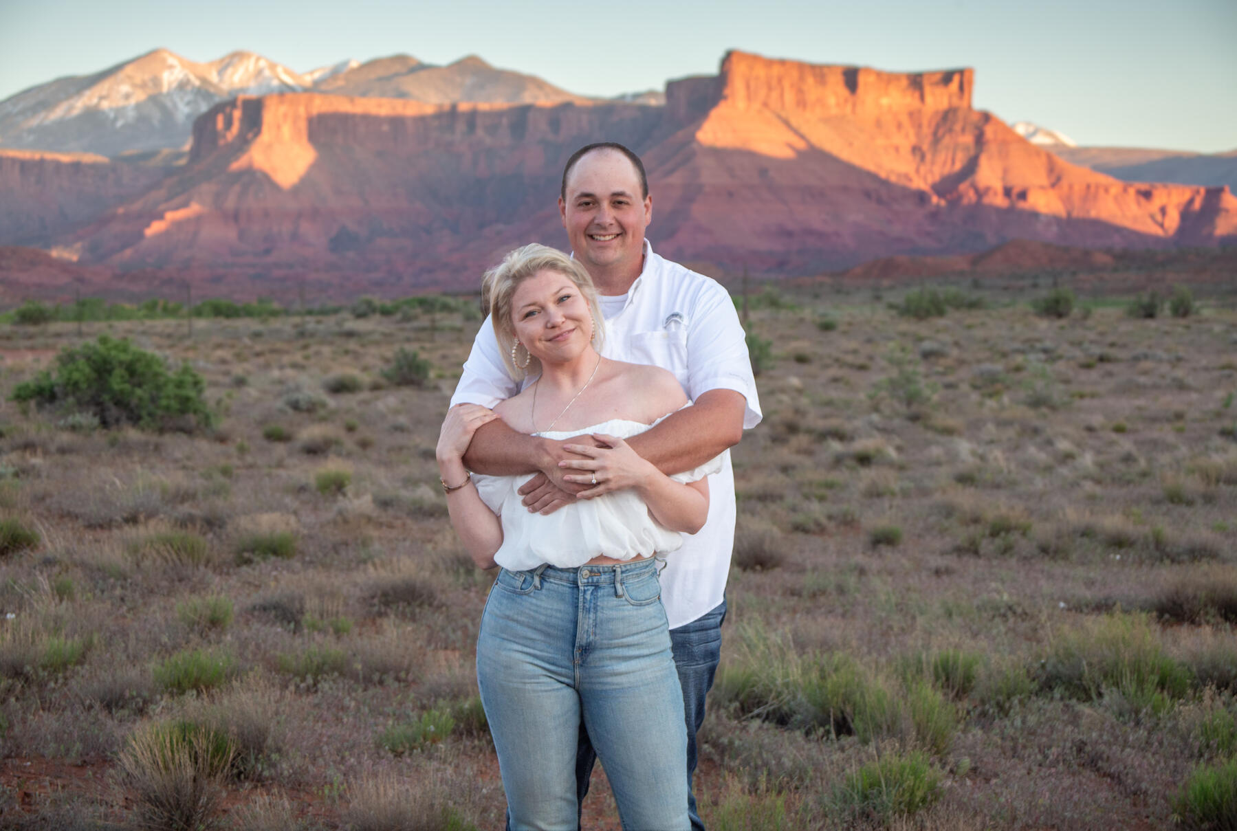 couple standing in front of red canyons and la sal mountains, soon to be husband standing behind with arms wrapped around soon t Moab proposal engagement